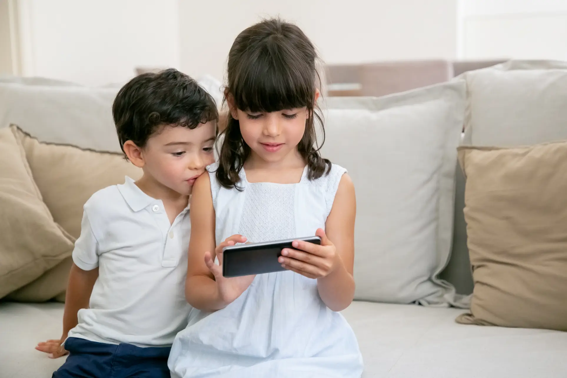 A girl holds a smartphone, while a boy leans in beside her on a cozy sofa.