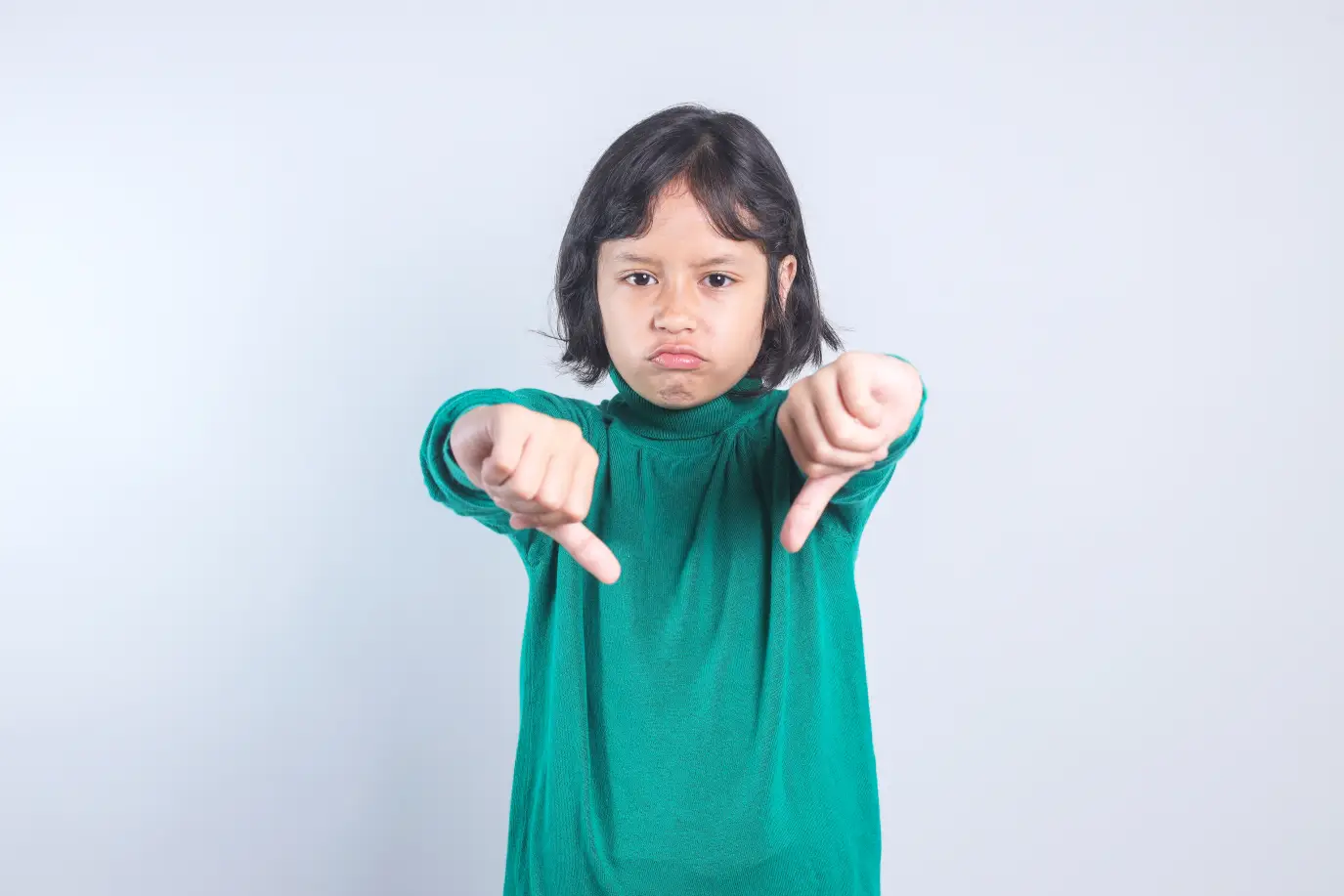 A child in a green sweater holds both hands down in a thumbs-down gesture against a light gray background.